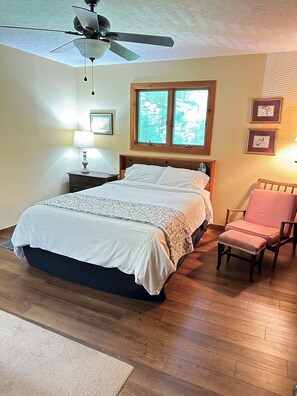 A bedroom with a bed, a chair, a ceiling fan, a window, and framed pictures on the wall.