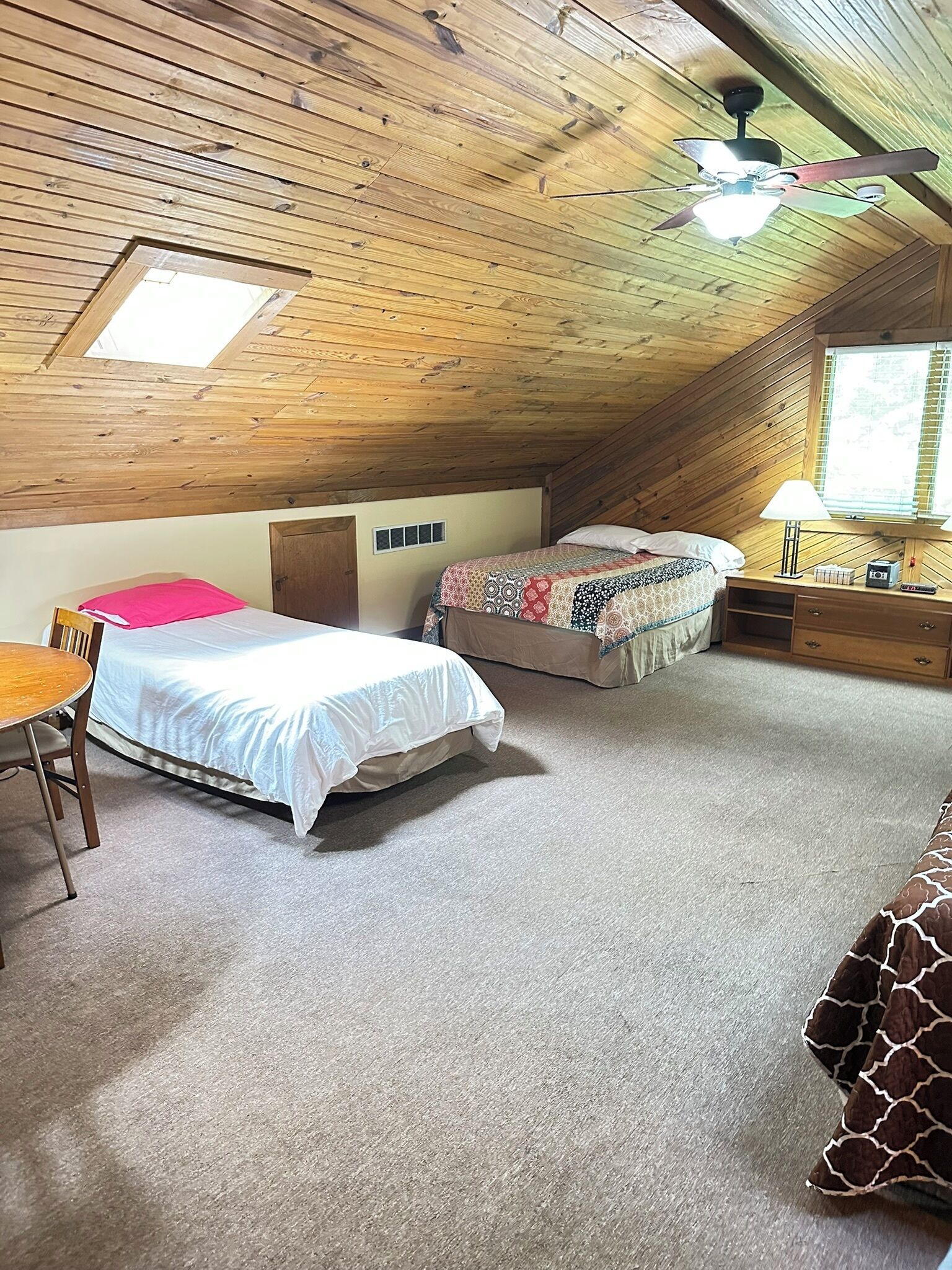 Ein Zimmer mit schrägem Dach, einem Bett mit weißer Bettdecke, einem kleineren Bett mit gemusterten Laken, einem Schreibtisch mit Lampe und einem Fenster mit Jalousien.