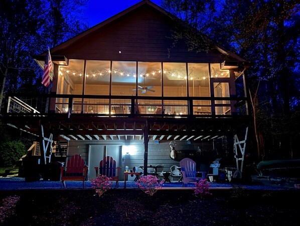 A two-story wooden cabin with large windows, a deck, and string lights.