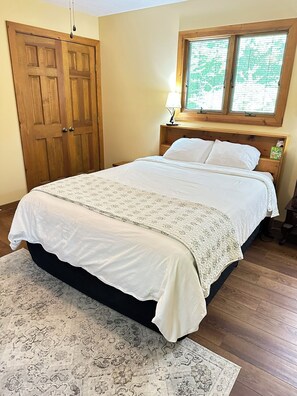 Un dormitorio con cama, una puerta de madera, una ventana con persianas y una mesita de noche.
