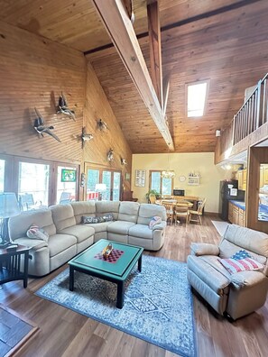 A spacious living room with a wooden ceiling, a sectional sofa, a coffee table, and a dining area with a chandelier.