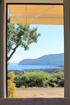A view from inside a room with a large window overlooking a coastal landscape, including a tree, hills, and a body of water. - Il Guercino (Capoliveri)