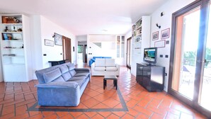 Un salon avec un canapé bleu, un fauteuil blanc et une petite table. On y trouve un téléviseur posé sur un meuble, ainsi qu’une bibliothèque remplie de livres et d’objets décoratifs.