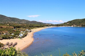 Une plage côtière aux eaux bleues et limpides, bordée d’un rivage sablonneux, avec des collines en arrière-plan.