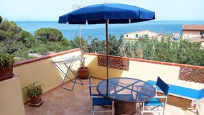A terrace with a round glass table and blue chairs, a blue umbrella, and potted plants. - La Piaggia (Rio)