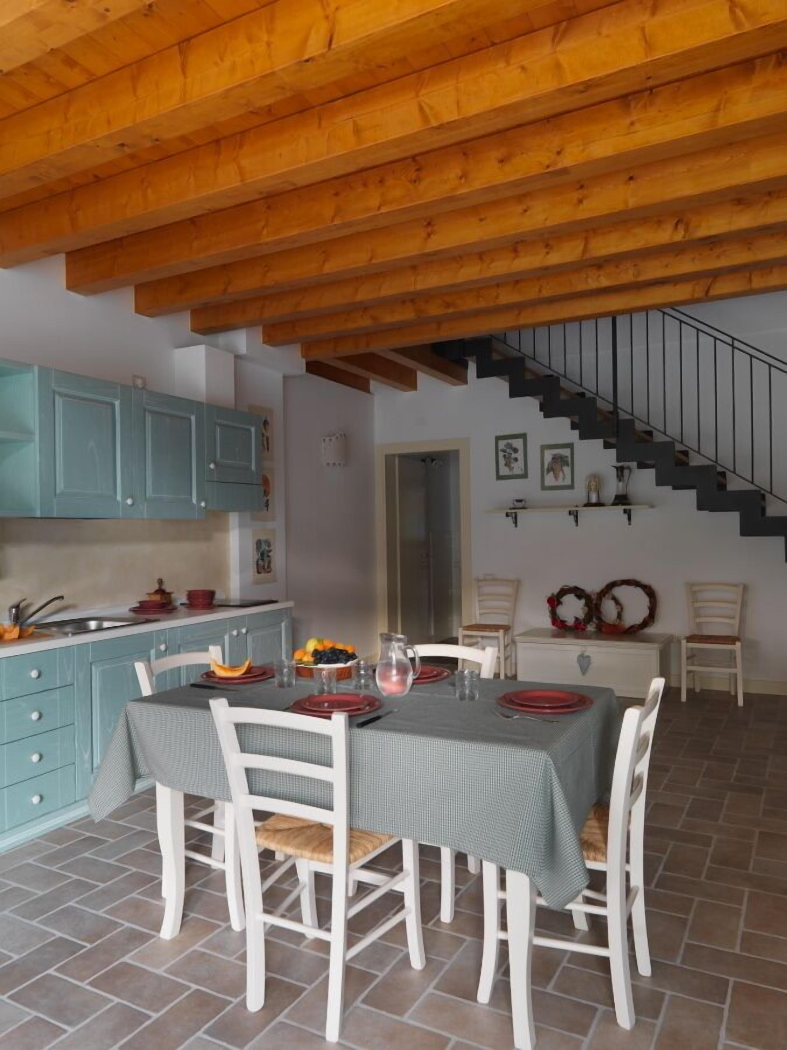 Un comedor con una mesa dispuesta para seis personas, vigas de madera en el techo, una escalera y una cocina con armarios color turquesa.