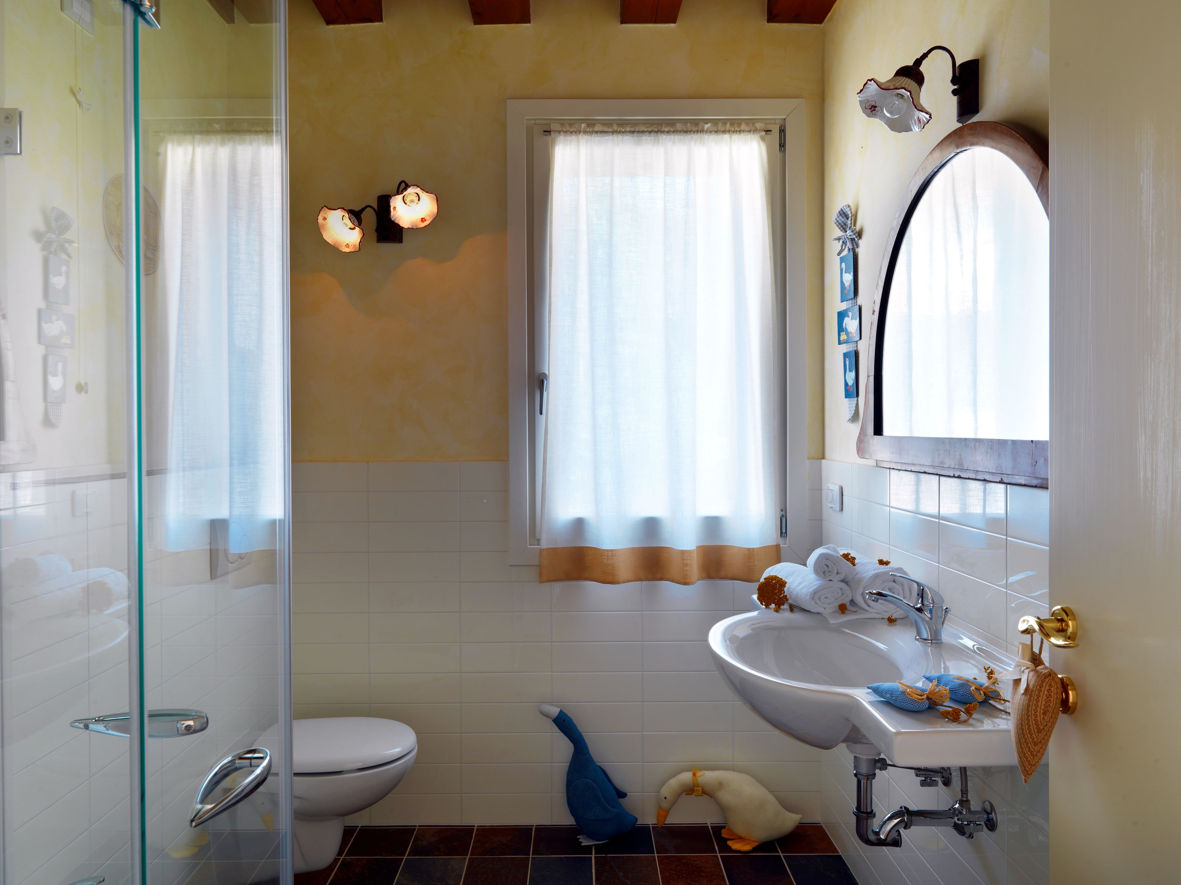 Ein Badezimmer mit Duschkabine aus Glas, einer weißen Toilette, einem Waschbecken mit Spiegel und einem Fenster mit Vorhängen.