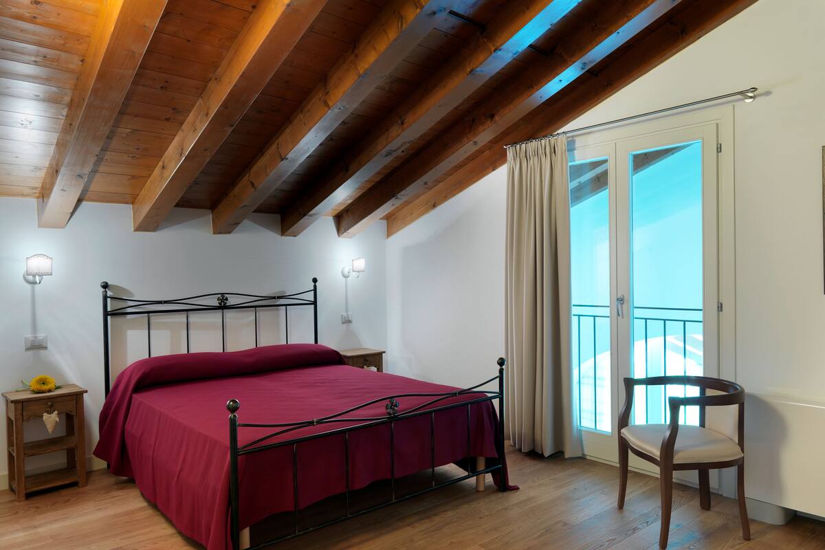 Un dormitorio con techo de madera, estructura de cama metálica, colcha roja, mesita de noche de madera con lámpara, una silla y una ventana con cortinas.