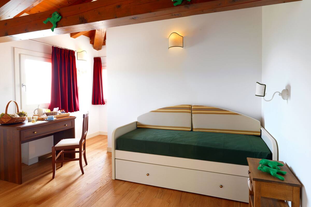 Un dormitorio con cama de madera, un escritorio y una silla. Hay una ventana con cortinas, una lámpara de pared y una mesita con un peluche verde.