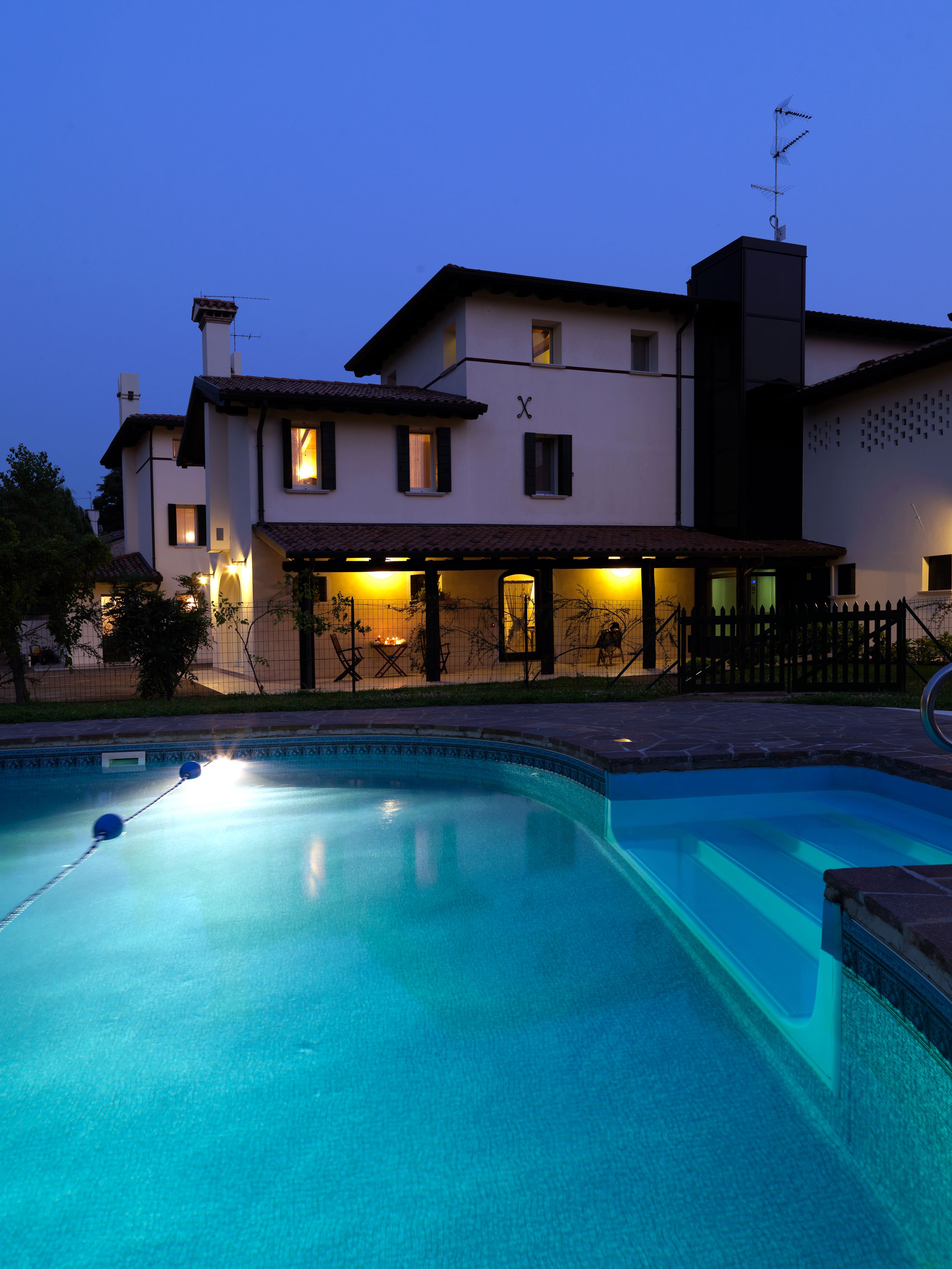A swimming pool with a tiled deck, a covered patio area, and a two-story building with multiple windows and a chimney.