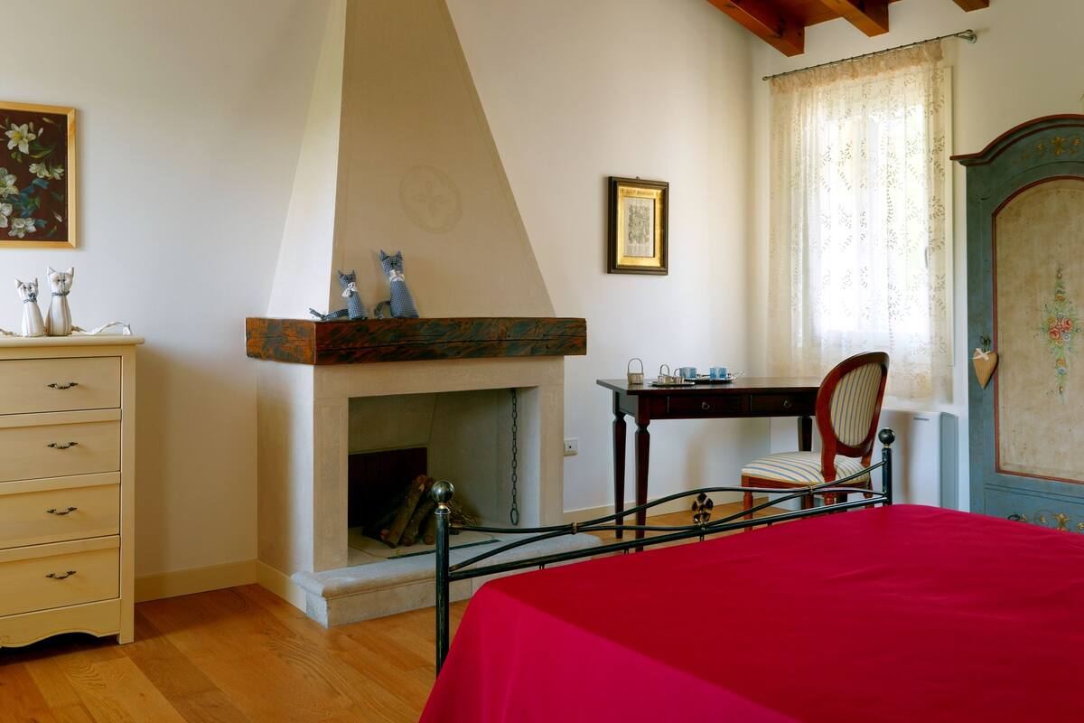 Un dormitorio con techo de madera, chimenea, cama con colcha roja, cómoda y una silla.