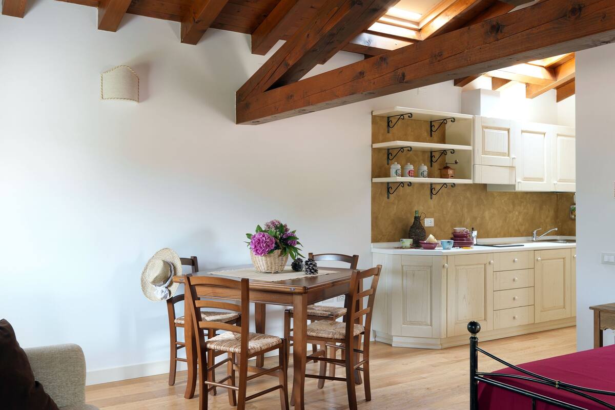 Un comedor con una mesa y sillas de madera, una cocina con armarios blancos y una cama con cubre cama rojo.