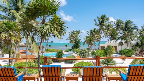 A wooden deck with four chairs and a table overlooking a beach and pool area, surrounded by palm trees and clear skies. - San Pedro 5-bedroom, 4-bathroom Villa Pearl, ocean views, concierge onsite (San Pedro)