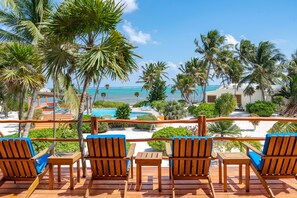 Une terrasse en bois avec quatre chaises et une table, donnant sur une plage et une piscine, le tout entouré de palmiers et sous un ciel dégagé.