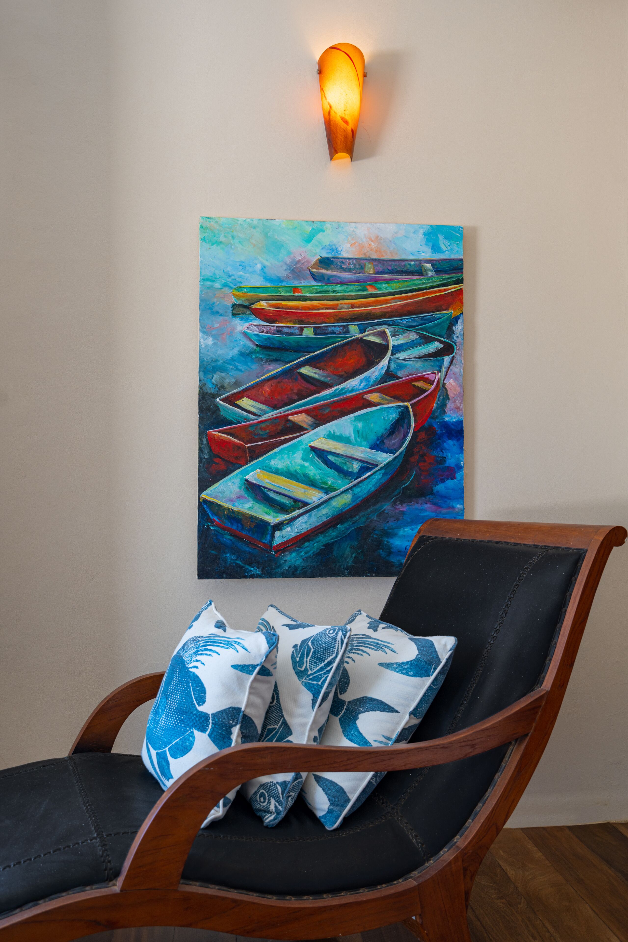 Een schilderij van kleurrijke boten op een muur, een houten stoel met kussens en een wandlamp.