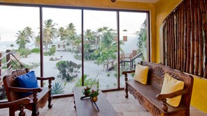 A room with a wooden bench and armchair, a small table with a vase, and large windows offering a view of a beach and palm trees. - San Pedro 5-bedroom, 4-bathroom Villa Pearl, ocean views, concierge onsite (San Pedro)