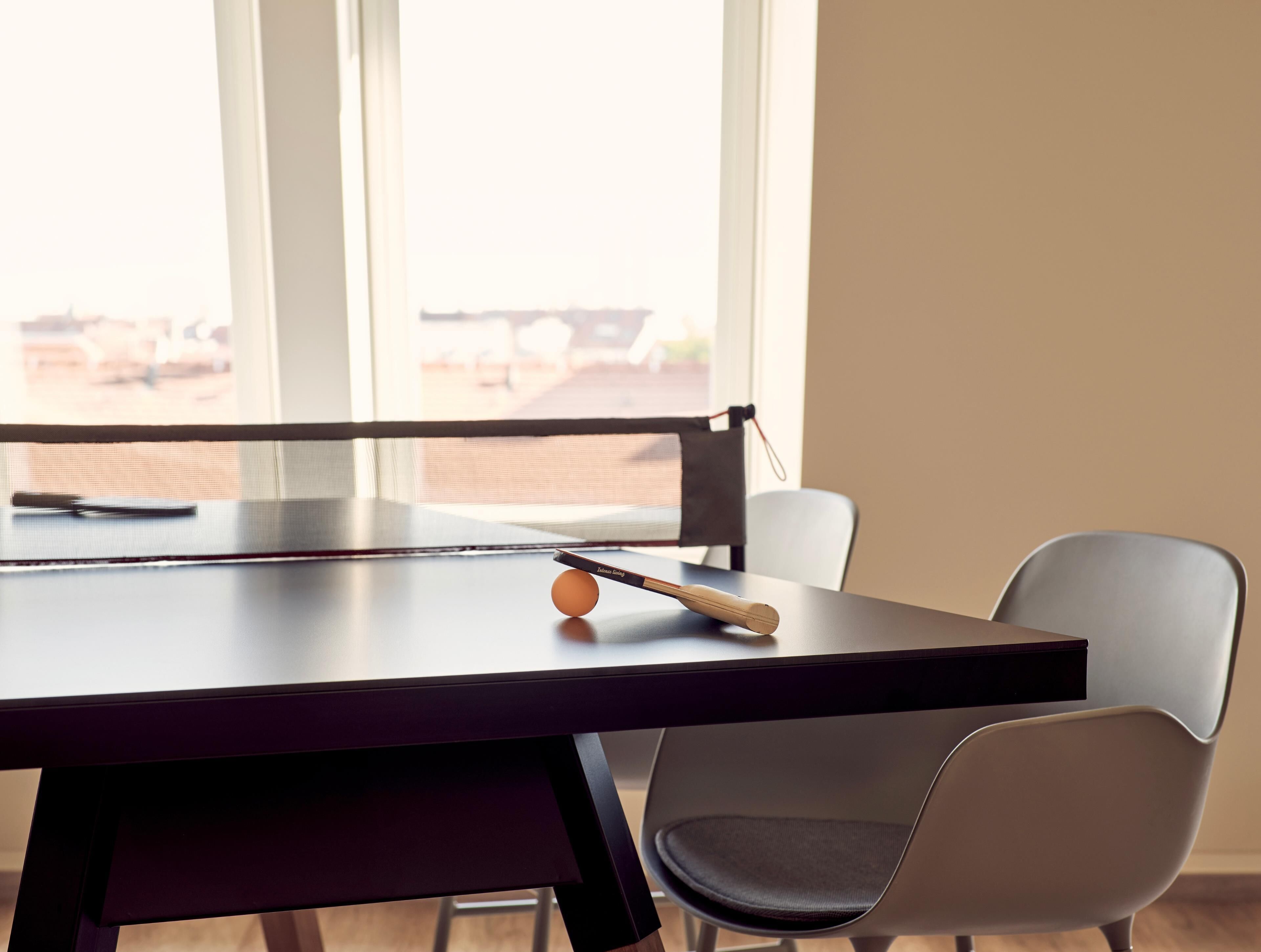 A table with a ping pong paddle and ball, a chair, and a window with a view of buildings.