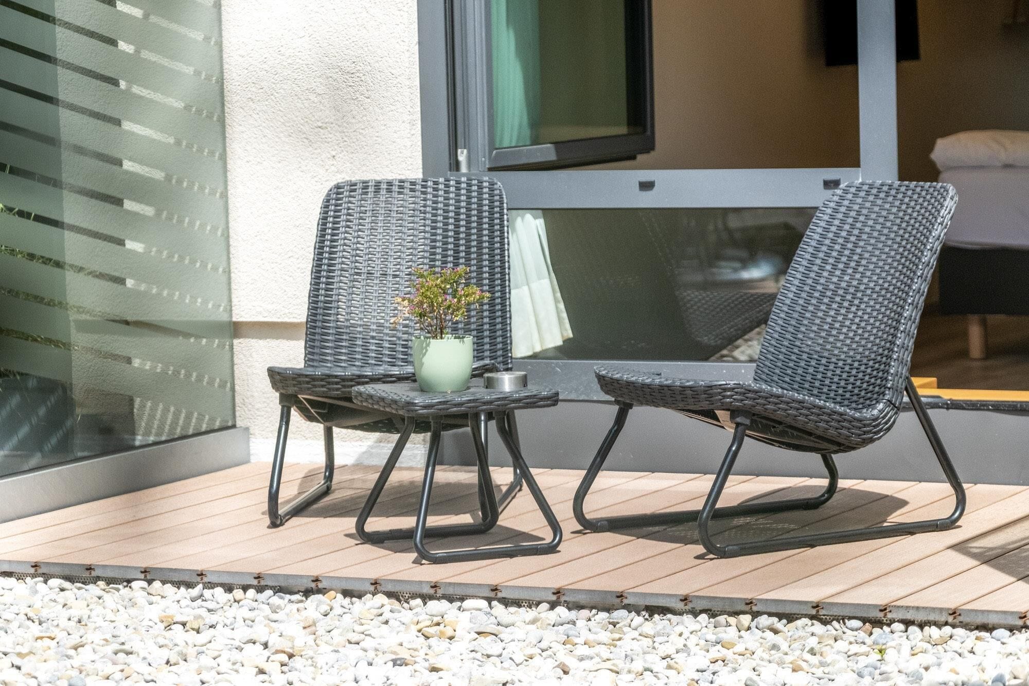 Zwei Korbstühle mit Metallgestellen auf einer Holzdielenveranda, ein kleiner Tisch mit Blumenvase und eine Glaswand mit Blick auf ein Gebäude.