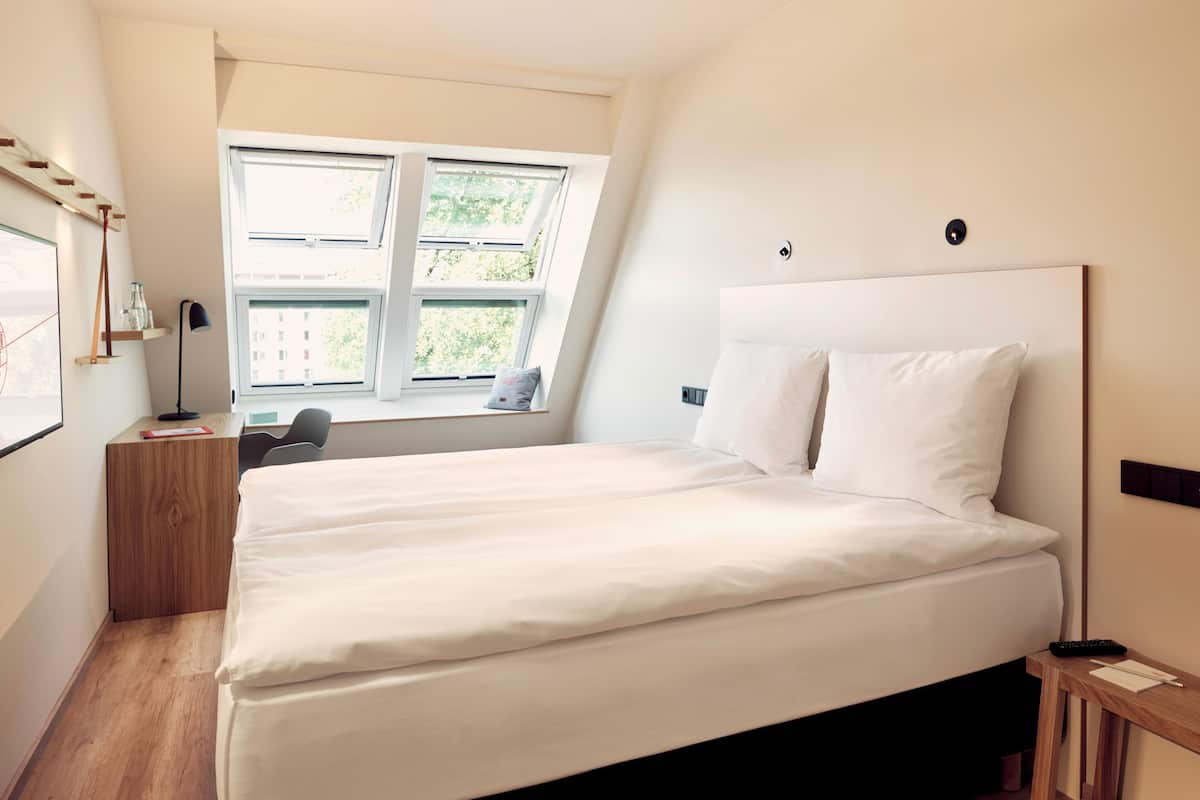 A modern bedroom with a large bed, a desk, and a window with a view of greenery.