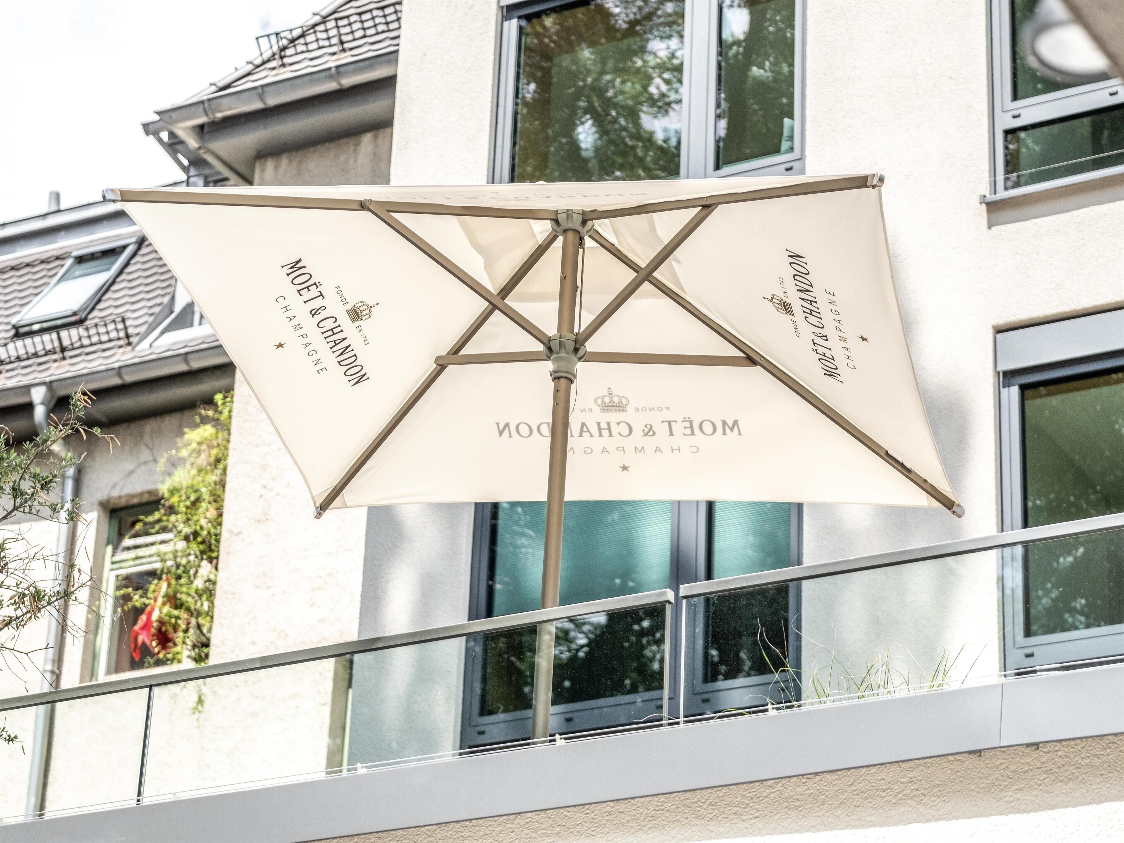 A large outdoor umbrella with 'MOËT & CHANDON' branding, mounted on a metal stand, positioned in front of a building with multiple windows and a balcony railing.