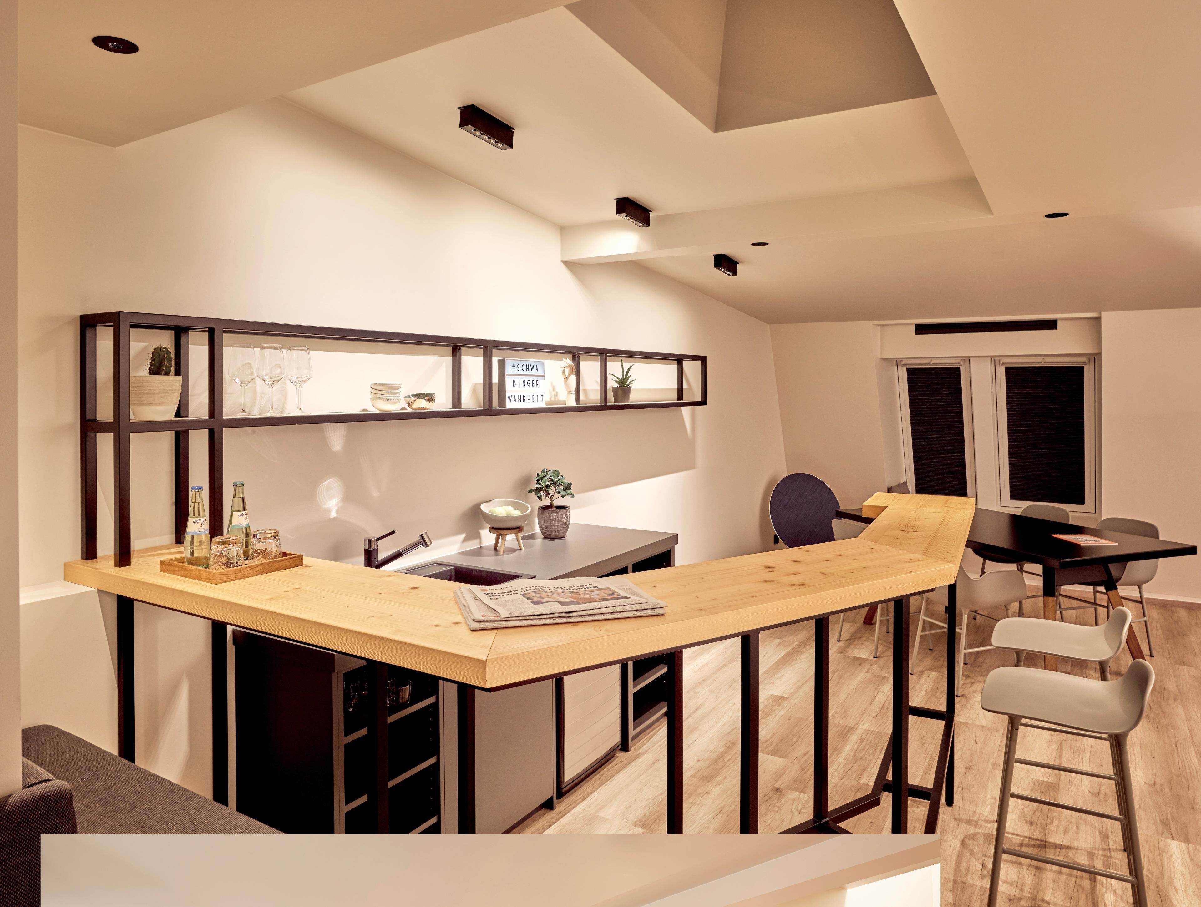 A modern kitchen with a wooden bar counter, a built-in shelf with decorative items, and a dining area with a black table and chairs.