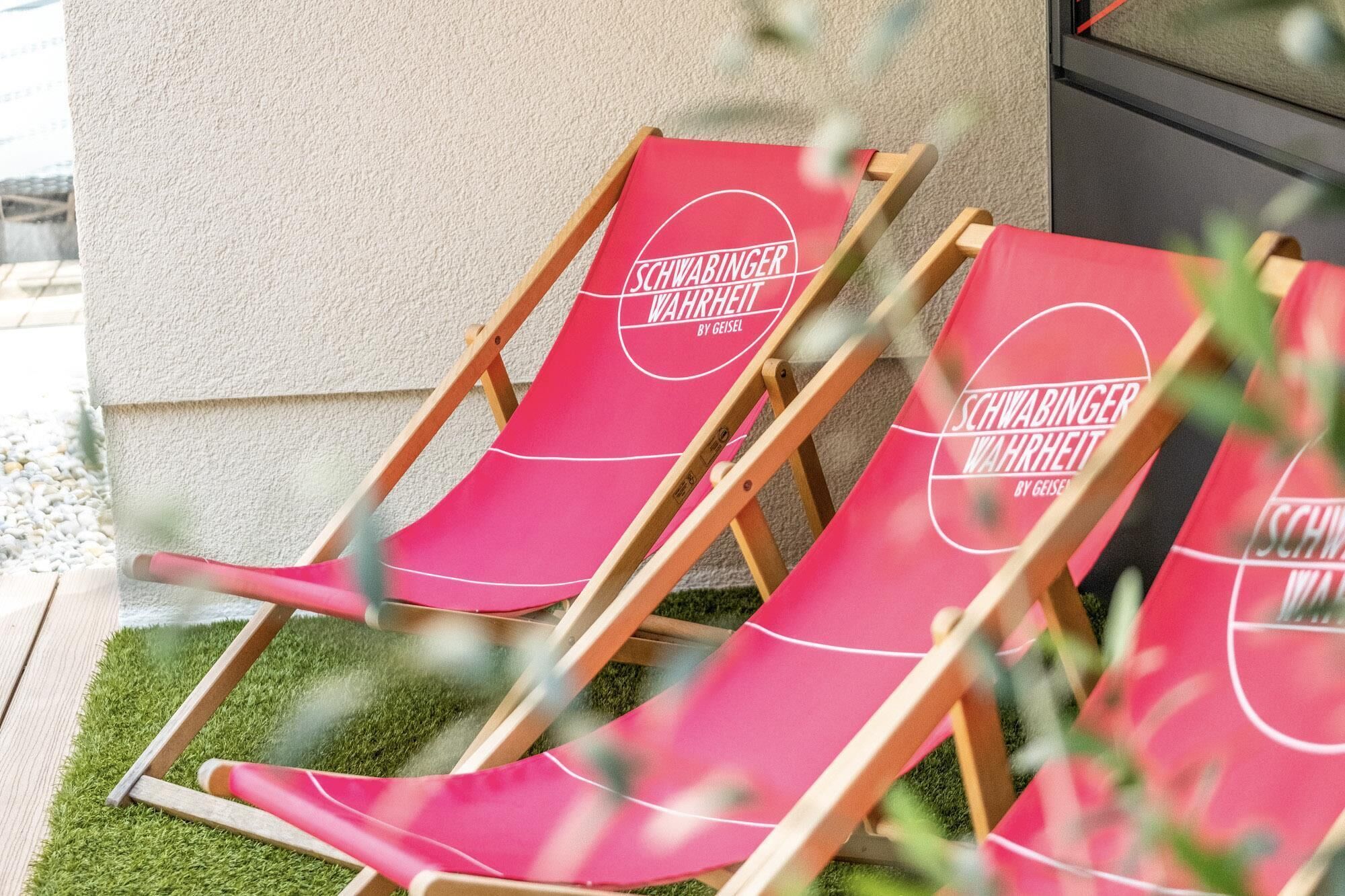 Two red lounge chairs with 'Schwabinger Wahrheit' branding, positioned against a wall with a small patch of grass.