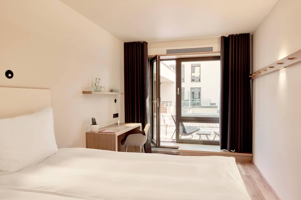 A hotel room with a bed, a desk, a chair, a small table with a plant, and a view of the outside balcony.