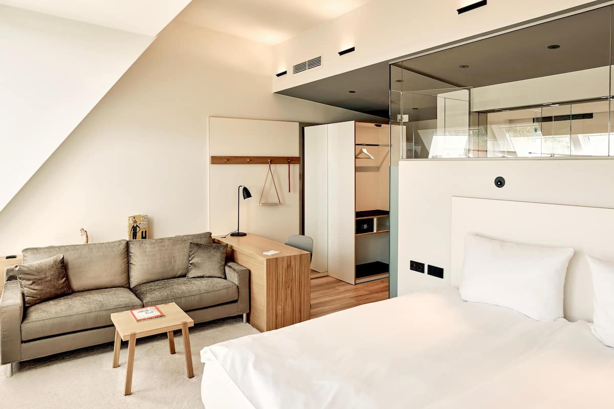 A modern hotel room with a sofa, a small table, a bed, a desk, and a walk-in closet.