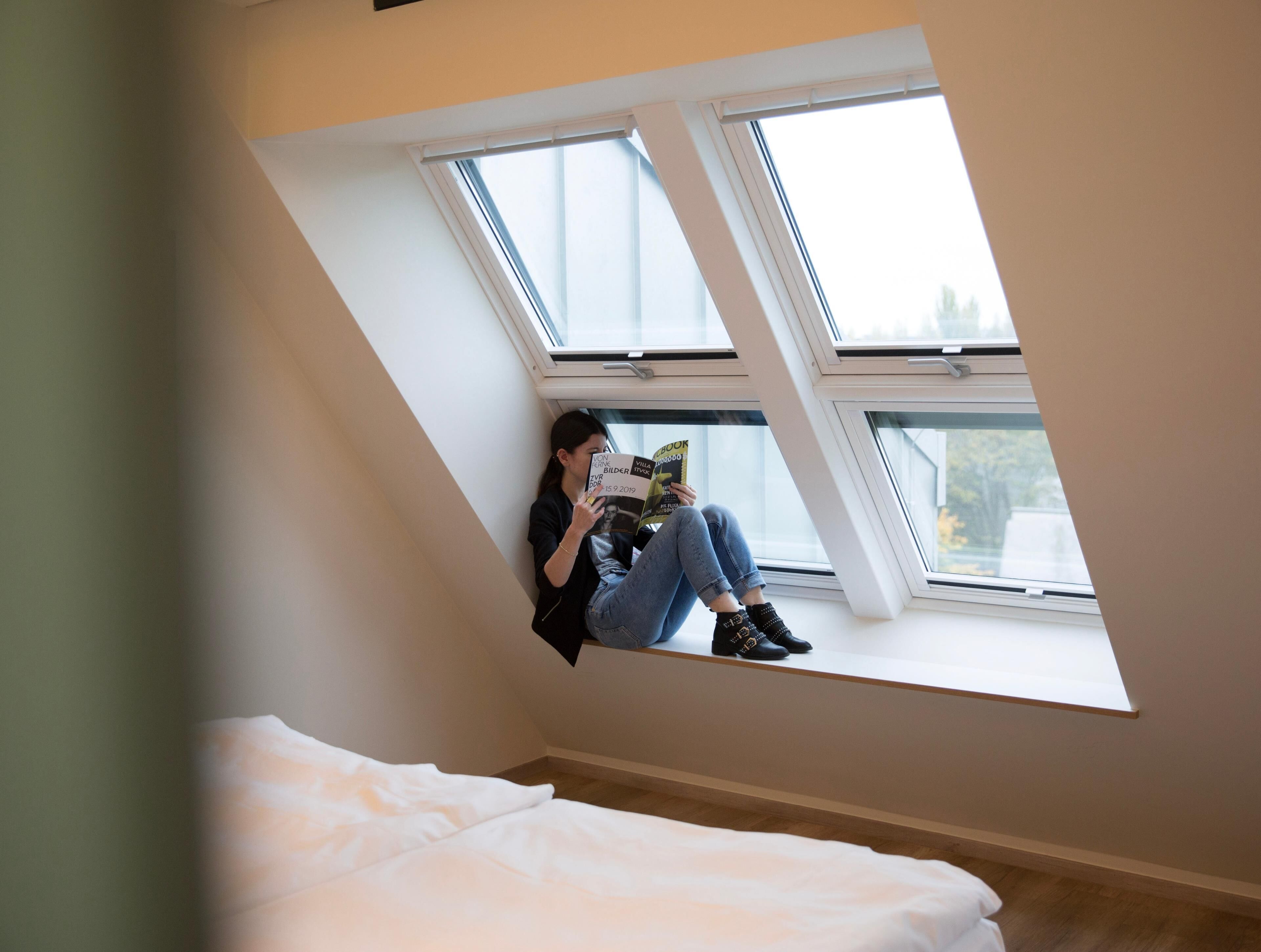 A person sitting on a window sill in a room with a sloped ceiling, reading a book.