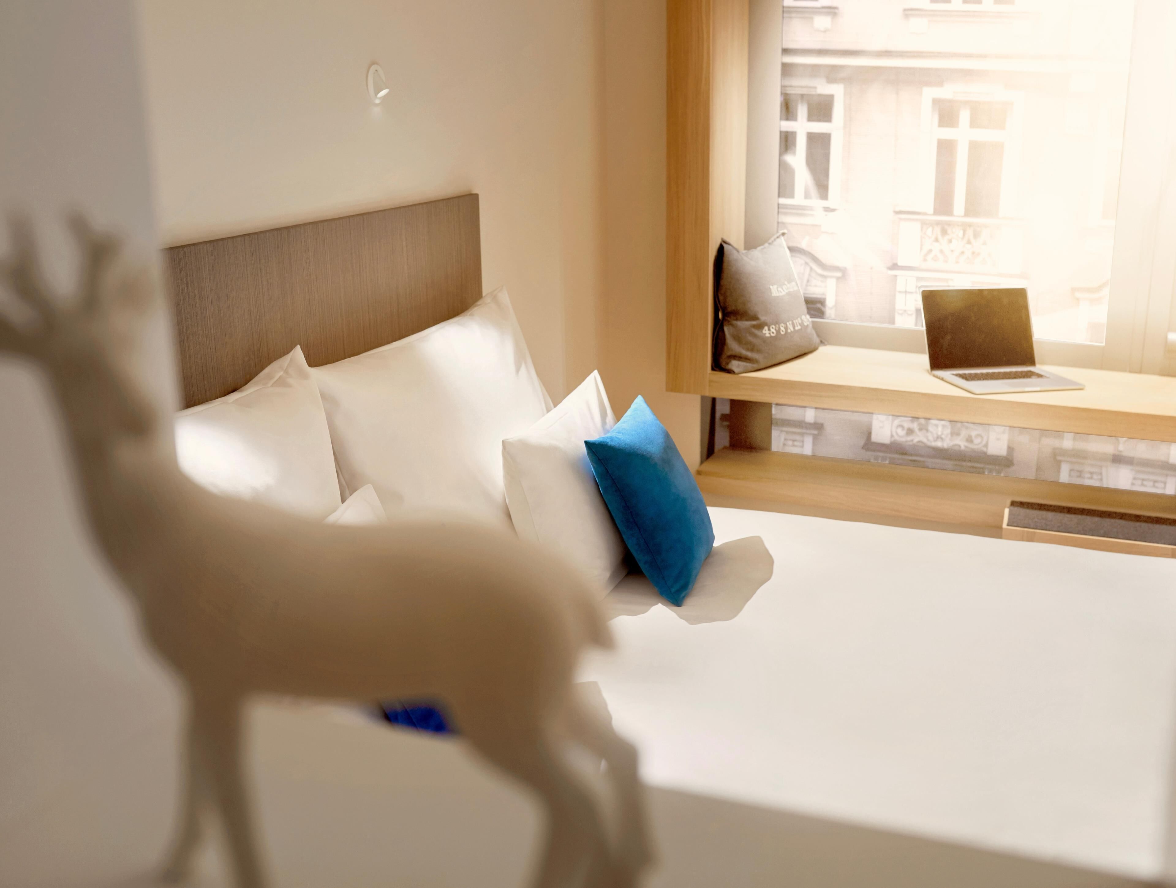A bed with white linens and a blue pillow, a laptop on a wooden shelf, and a window with a view of buildings.