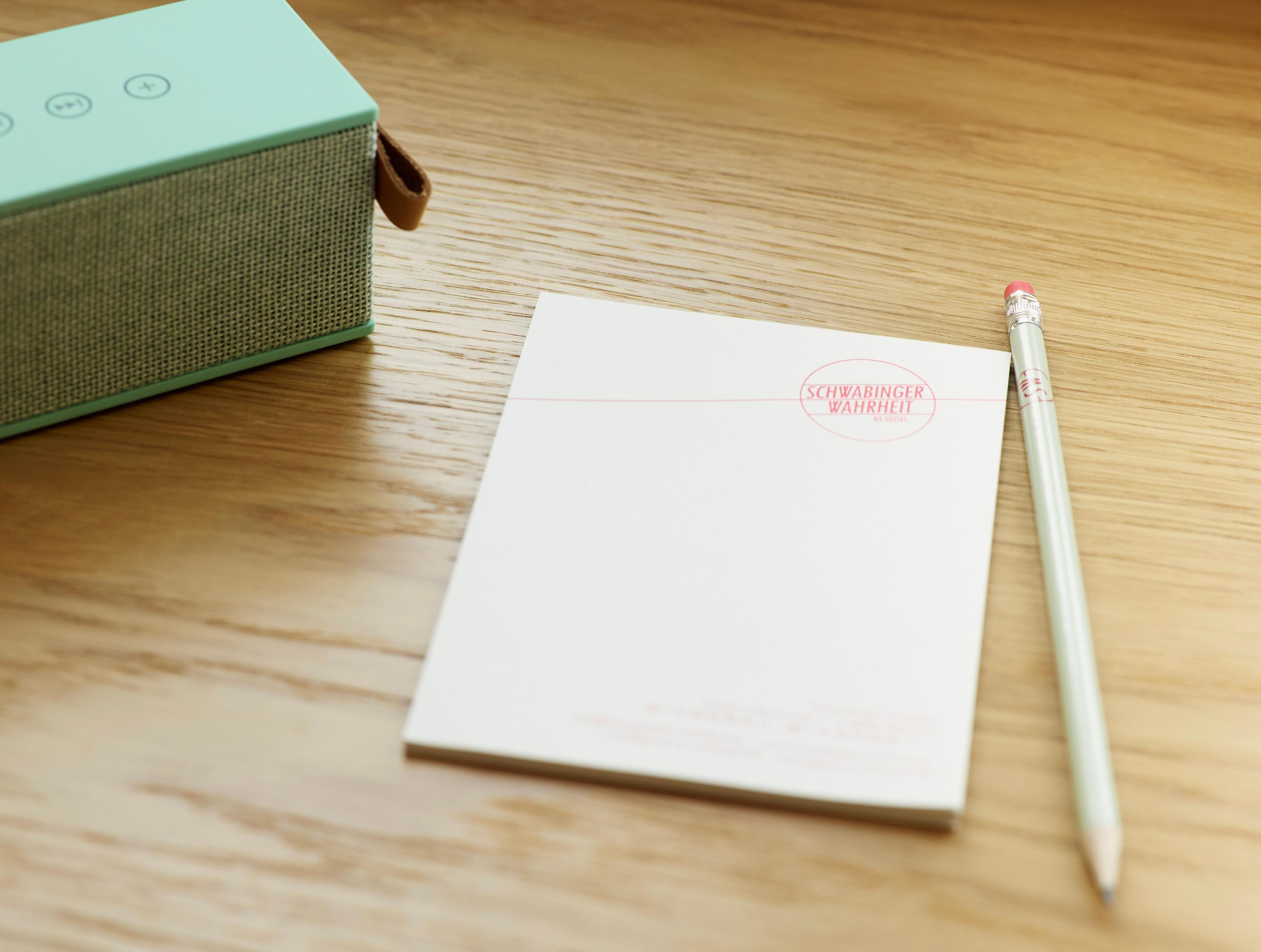 A wooden surface with a white notepad, a pencil, and a green speaker.