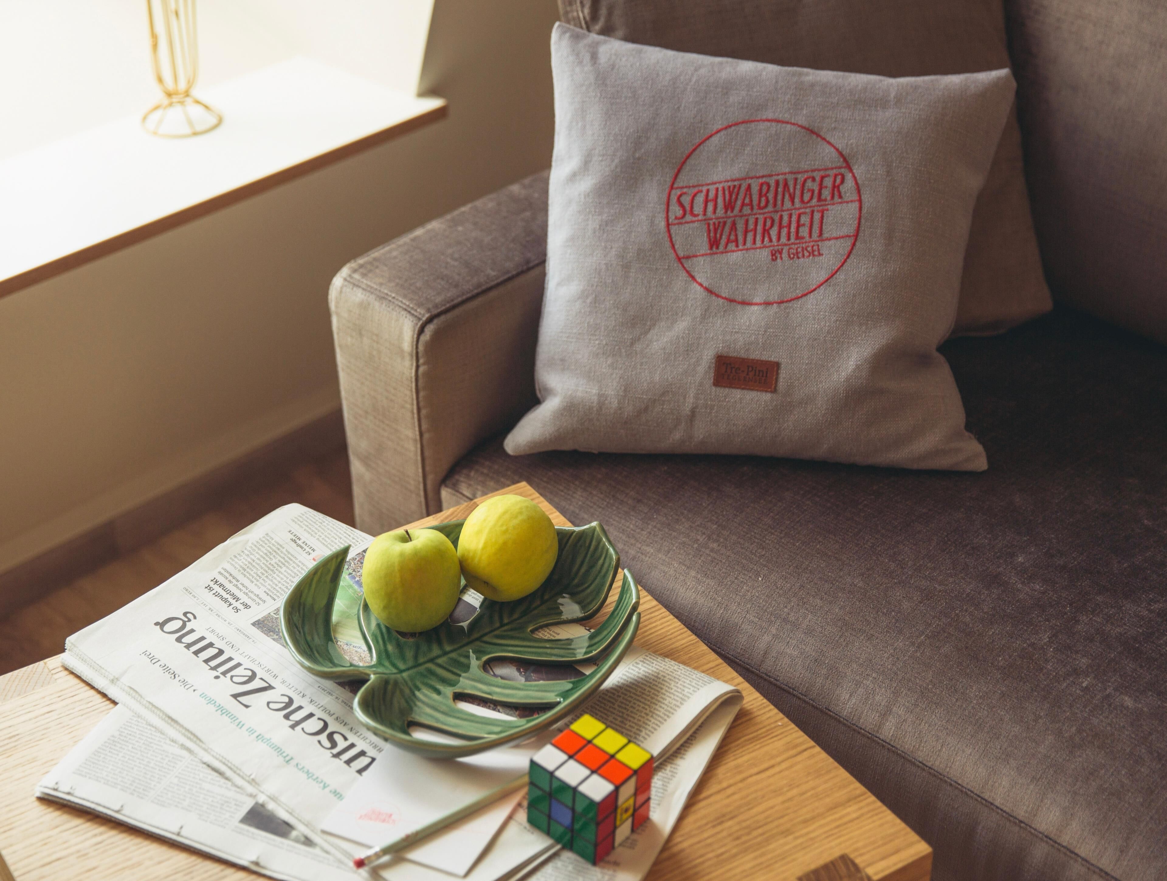 Una sala de estar acogedora con un sofá gris, un cojín con un logo, una mesita de madera, un cuenco con limones, un cubo de Rubik y periódicos.