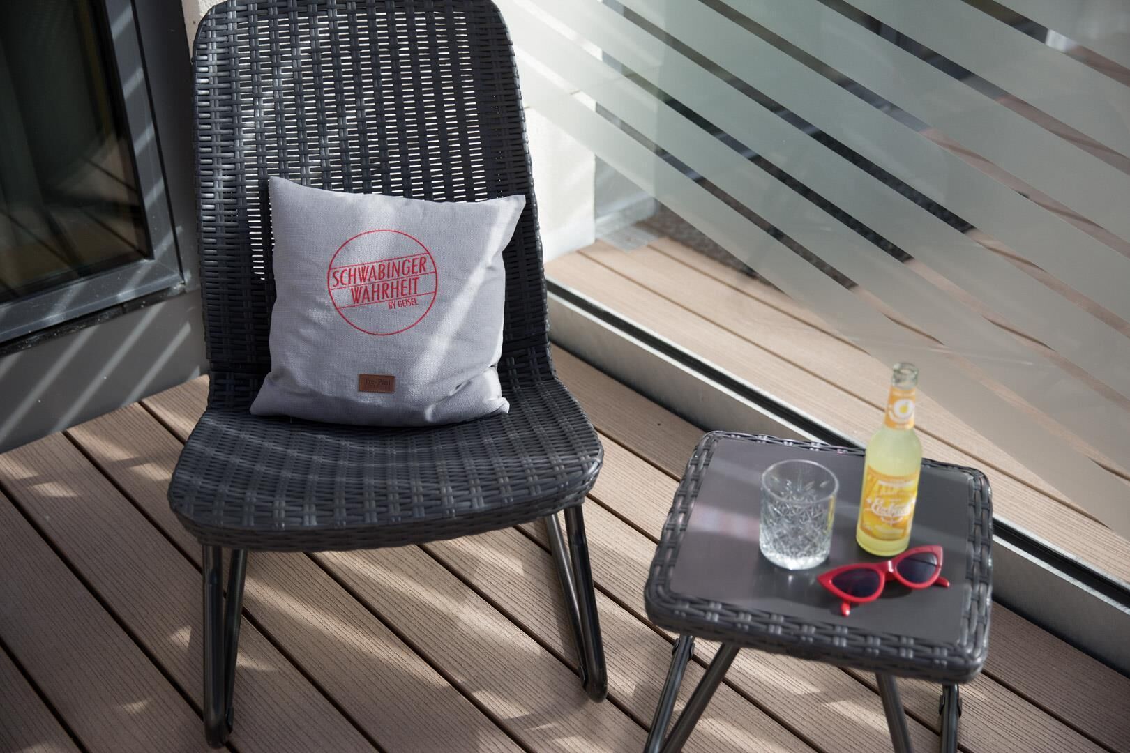 A wicker chair with a pillow, a small table with a bottle, a glass, and sunglasses.