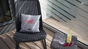 A wicker chair with a pillow, a small table with a bottle, a glass, and sunglasses.