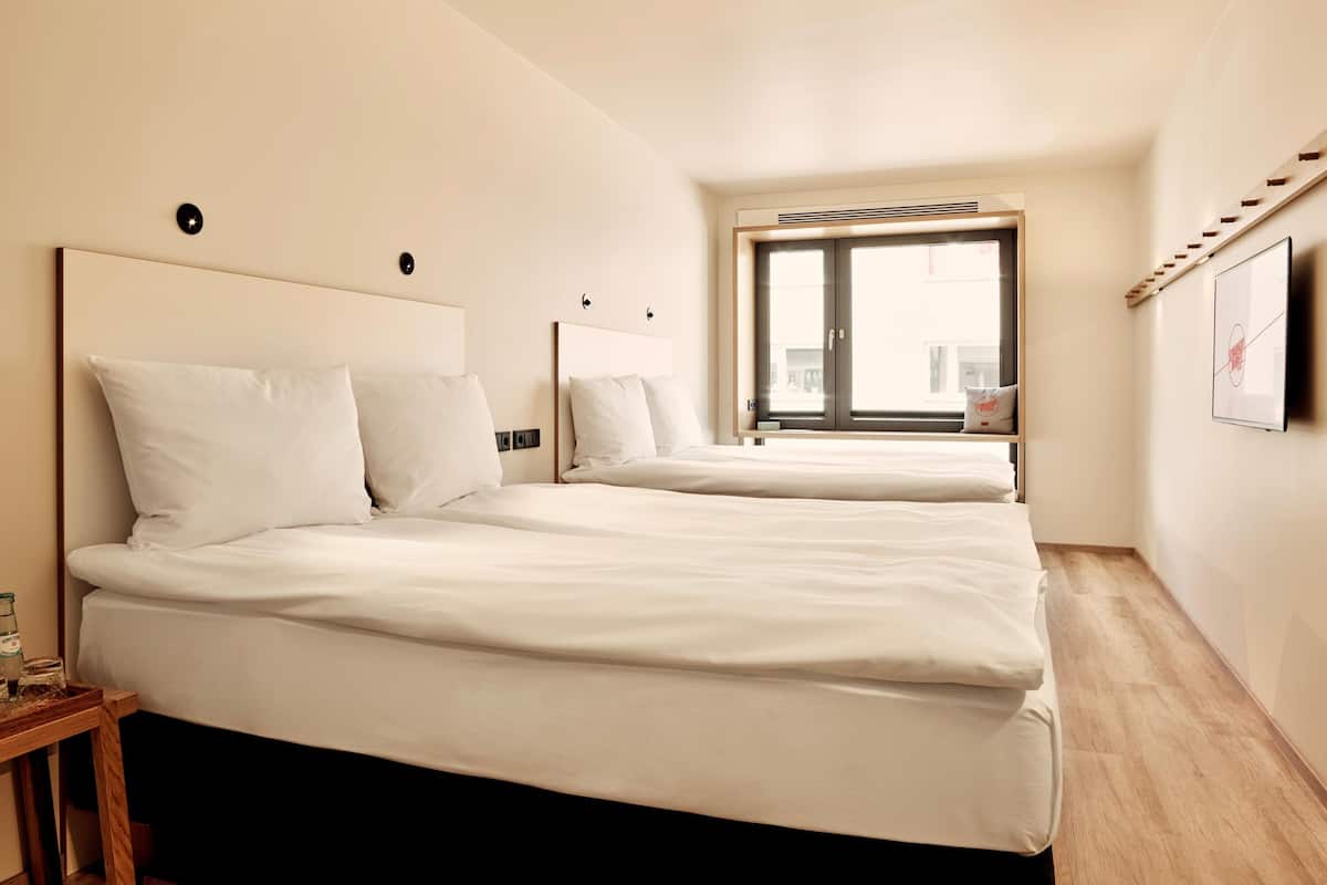 A hotel room with two beds, a window, a wooden floor, and a small table with a bottle and glasses.