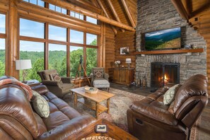 Interior de una cabaña de troncos con chimenea de piedra, sofás de cuero, televisor y un ventanal con vistas a un bosque.