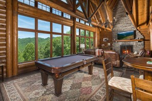 A log cabin interior with a pool table, leather chairs, a fireplace, and a large window overlooking a forest.