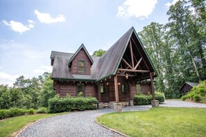 Cozy cabin with a gabled roof and porch, surrounded by lush greenery and a gravel driveway.