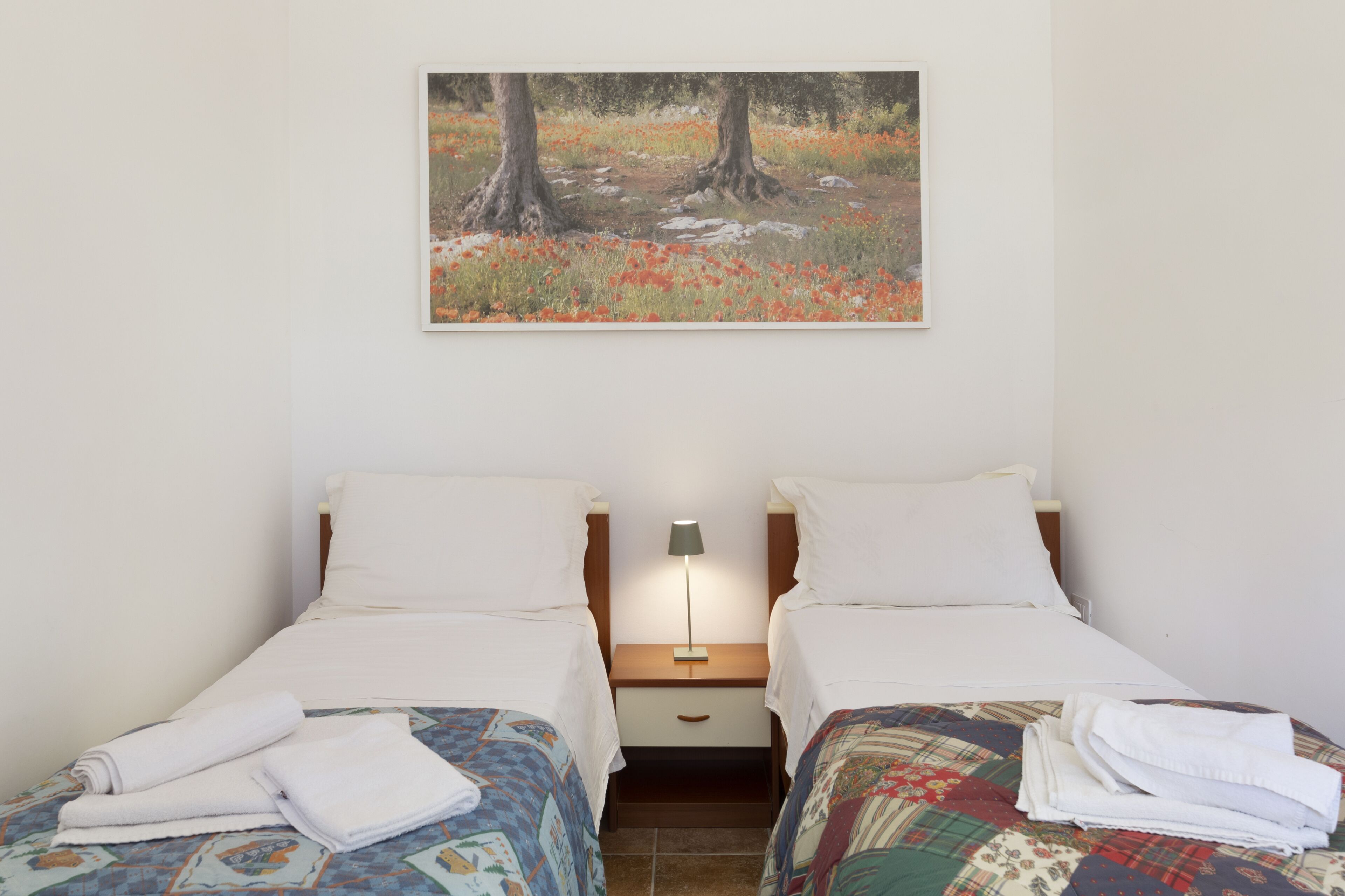 A room with two beds, a nightstand with a lamp, and a painting of a forest scene on the wall.