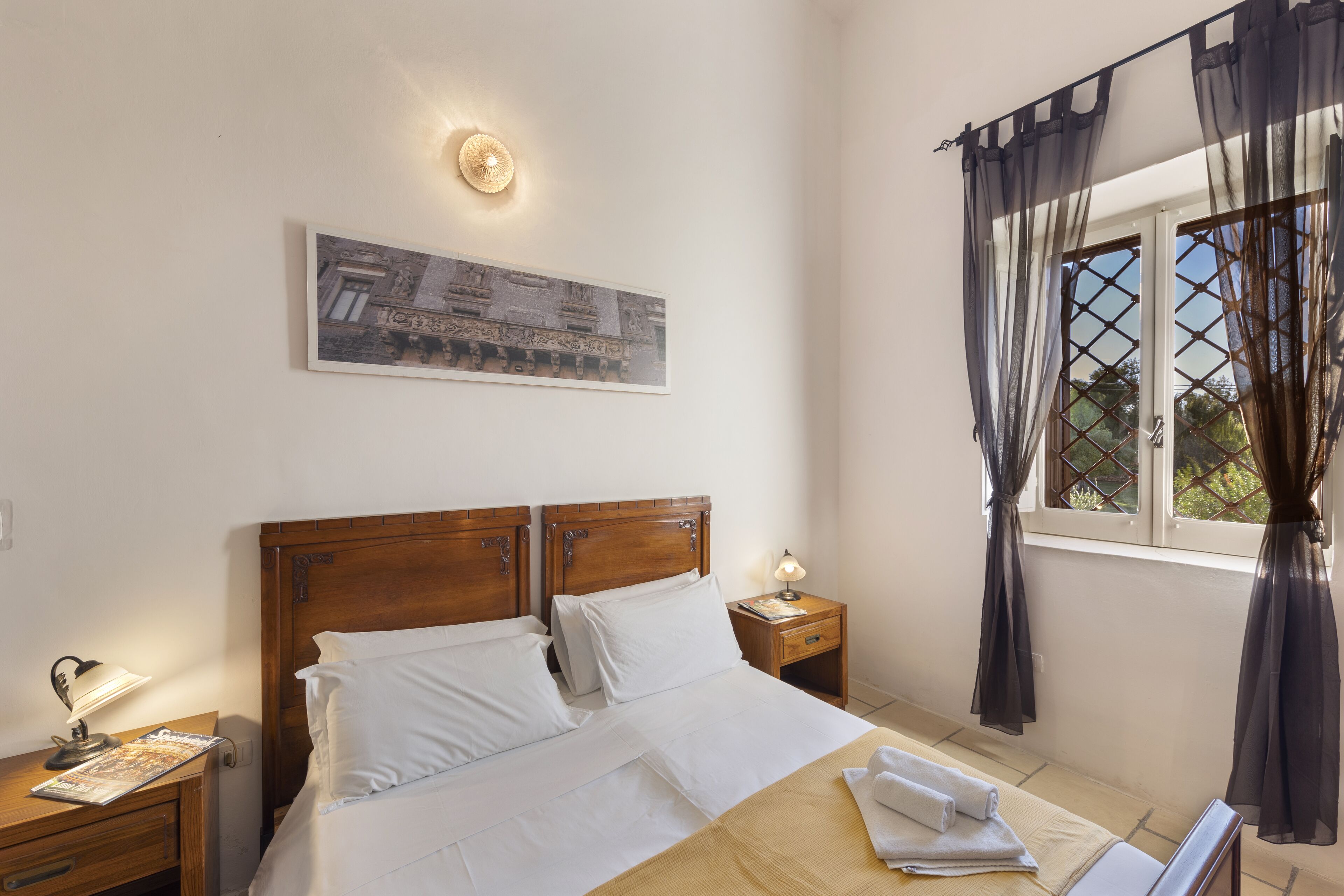 Un dormitorio ordenado con una cama de madera, una mesita de noche con lámpara, un cuadro en la pared y una ventana con cortinas.