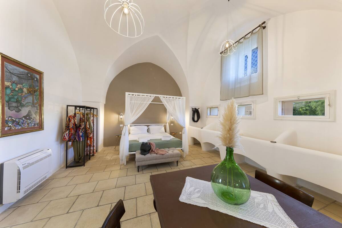 A bedroom with a bed, a bedside table with a vase, a chair, a sofa, a window, and a wall-mounted TV.