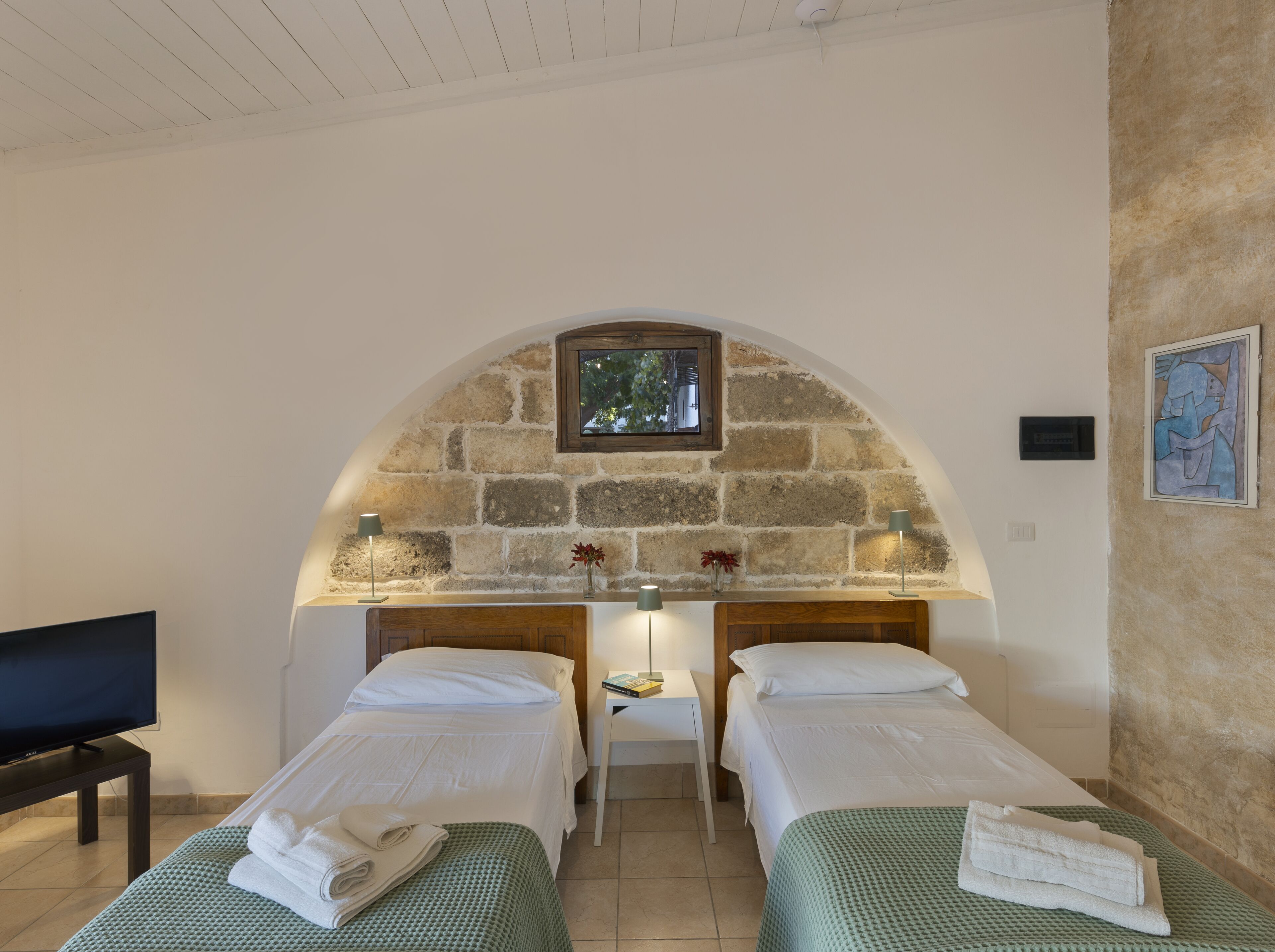 Una habitación con dos camas individuales, una mesita pequeña, un televisor y un arco de piedra con una ventana.