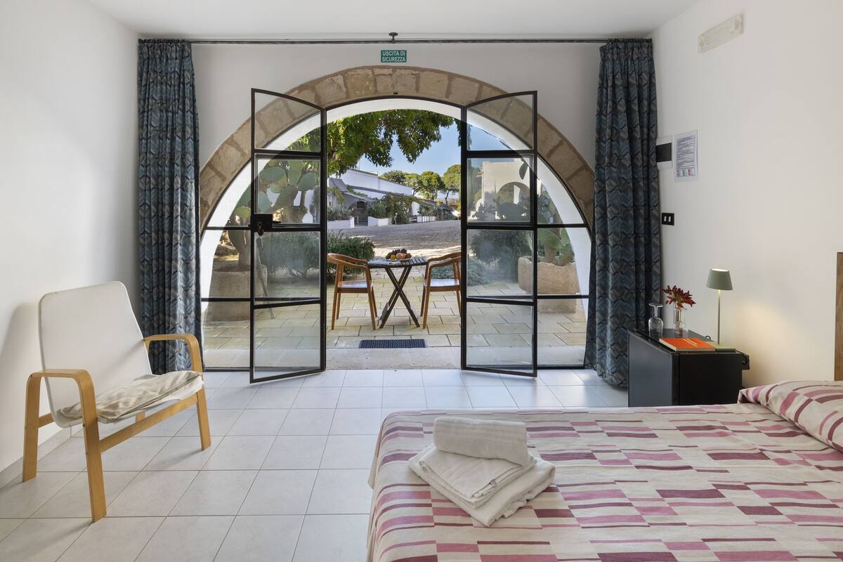 A hotel room with a bed, a chair, a desk with a lamp, and a view through a glass door to an outdoor area with a table and chairs.