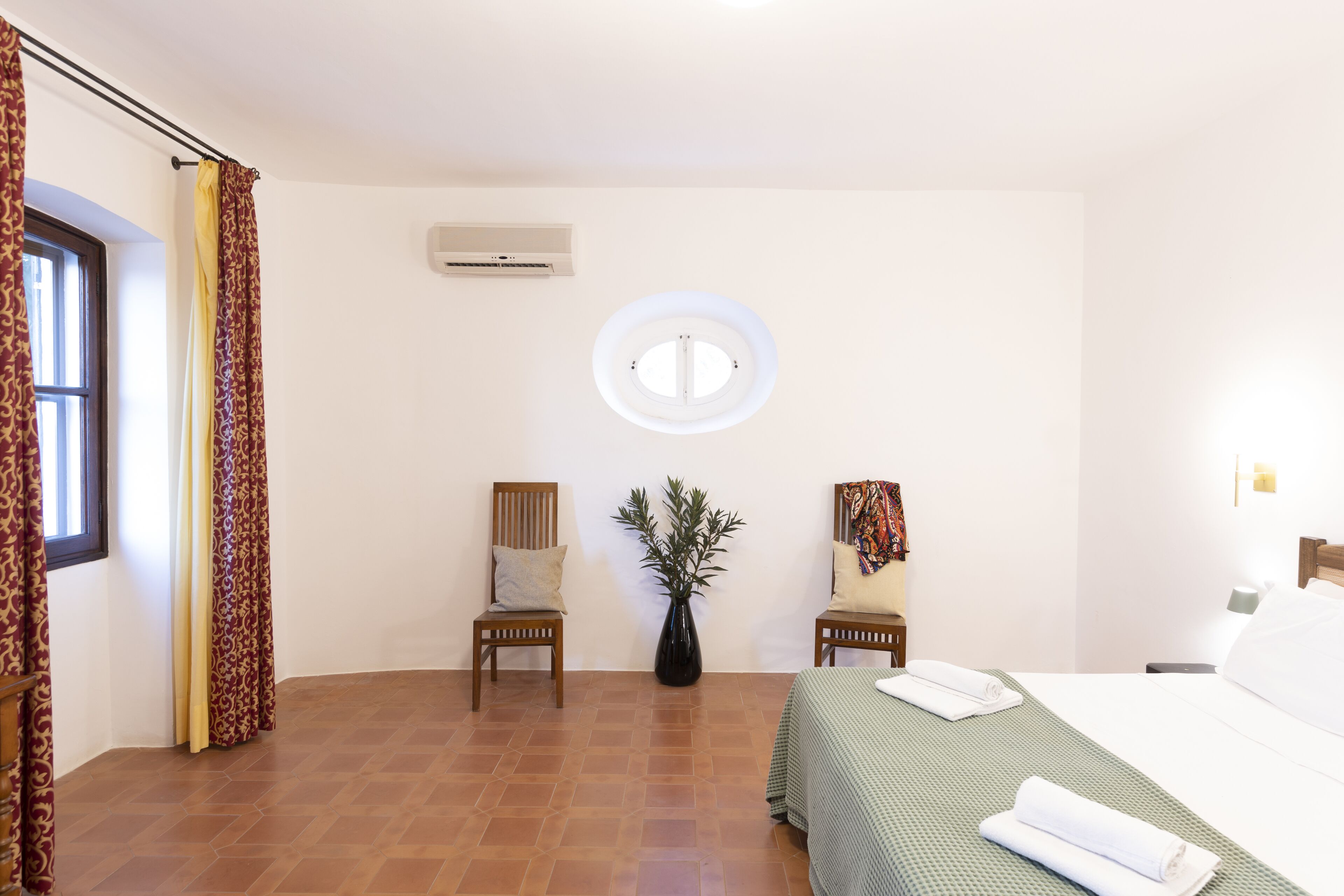 A hotel room with a bed, two chairs, a potted plant, and a window with curtains.