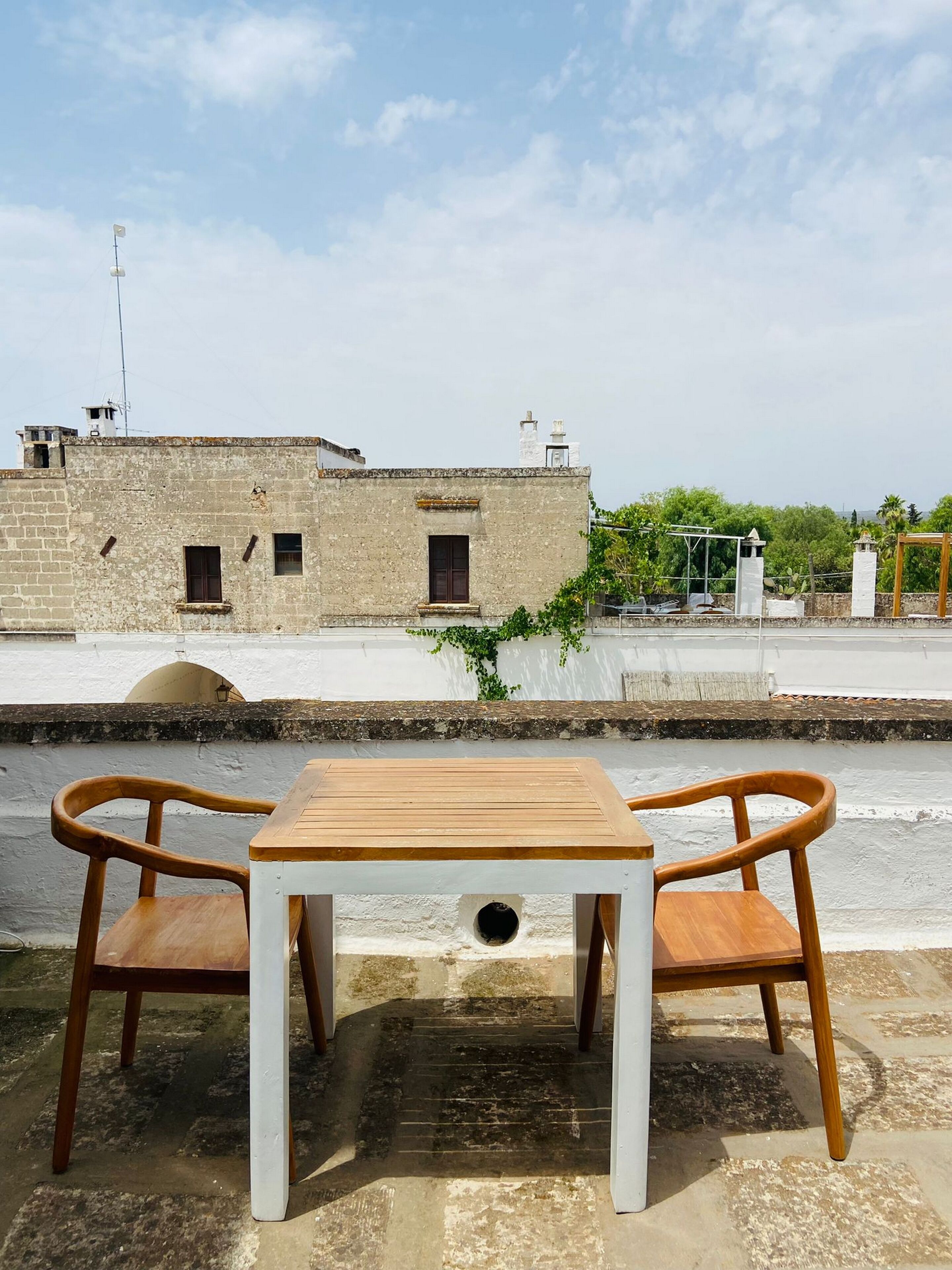 A rooftop terrace with a wooden table and two chairs, overlooking a historic building with multiple windows and a white wall.