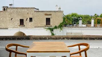 A rooftop terrace with a wooden table and two chairs, overlooking a historic building with multiple windows and a white wall.
