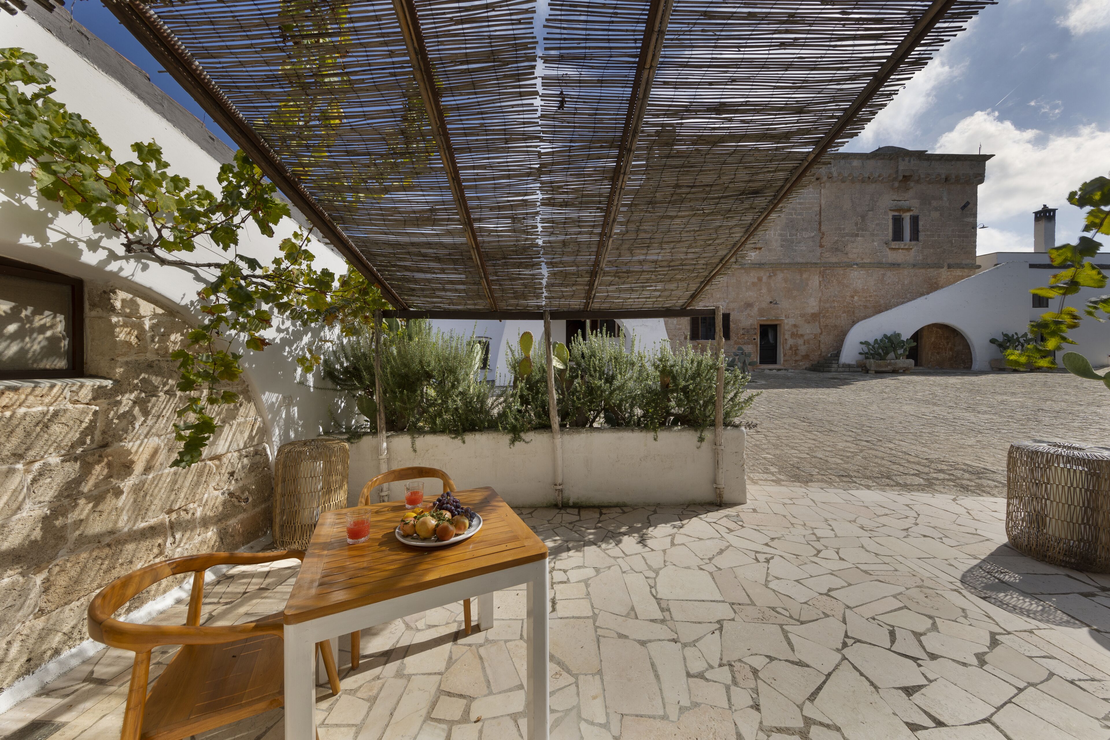 An outdoor patio with a wooden table set for two, surrounded by stone flooring and a pergola overhead.