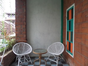 Pequeño patio con dos sillas blancas de mimbre, una mesa redonda, pared de ladrillo y una ventana con detalles verdes.