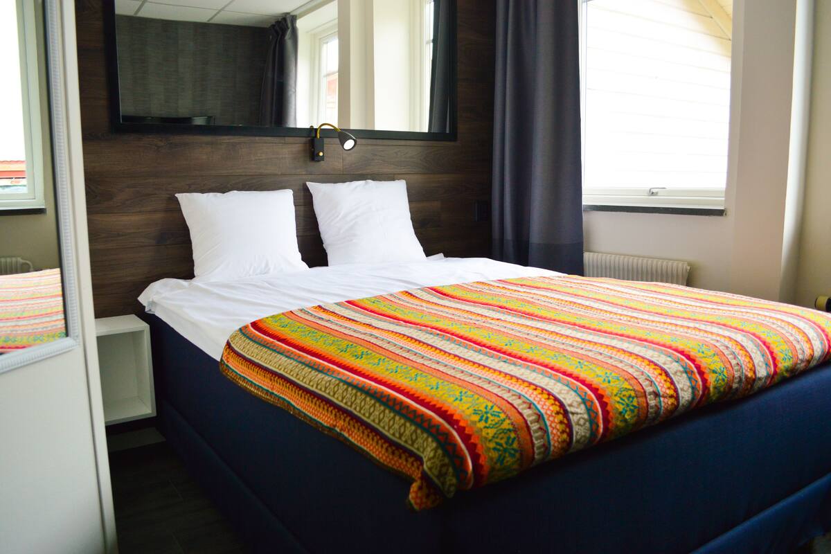 Ein ordentlich bezogenes Bett mit bunter Decke, ein Nachttisch, ein Spiegel über dem Bett und ein Fenster mit Vorhängen.