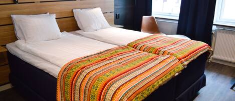 A double bed with a white duvet and two pillows, a patterned blanket, and a wooden headboard with two wall-mounted lights.